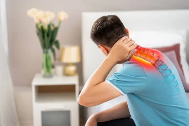 Hombre joven sentado en una silla sosteniendo la nuca con expresión de molestia por rigidez muscular causada por Dolor de cuello después de largas horas de trabajo.
